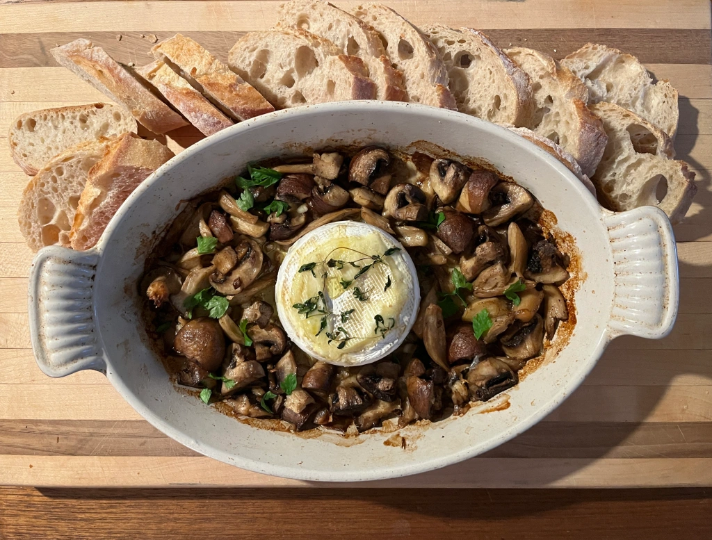 Photo of a serving dish containing roasted mushrooms with a round of cheese in the centre, placed on a cutting board surrounded by slices of baguette.